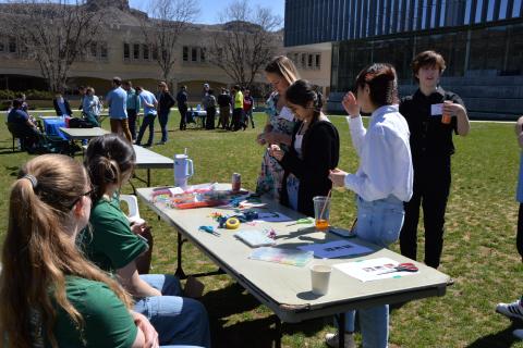 people mingle at tables set on grass near college buildings