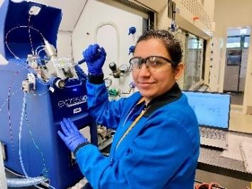 image of person wearing a blue lab coat, with one hand on handle of equipment in laboratory, CMI postdoc Sandeep Kaur, Oak Ridge National Laboratory