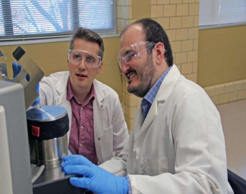 image of two people wearing laboratory coats: Ihor Hlova (back) and Tyler Del Rose (front) placing the ball milling chamber into the machine for processing.