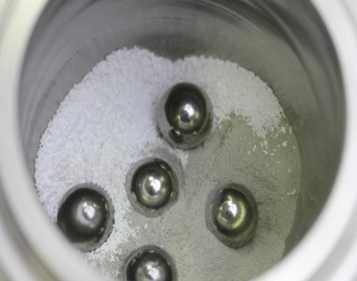 image of five metal balls in a cup with powder: Ball milling chamber contents: spodumene, reactant, and steel balls.