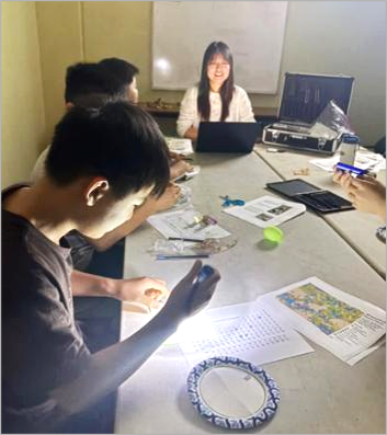 people seated at a table: CMI Leadership Academy member Eunjeong Kim, Lawrence Livermore National Laboratory (shown at top) guides students through experience with phosphor paper and light.