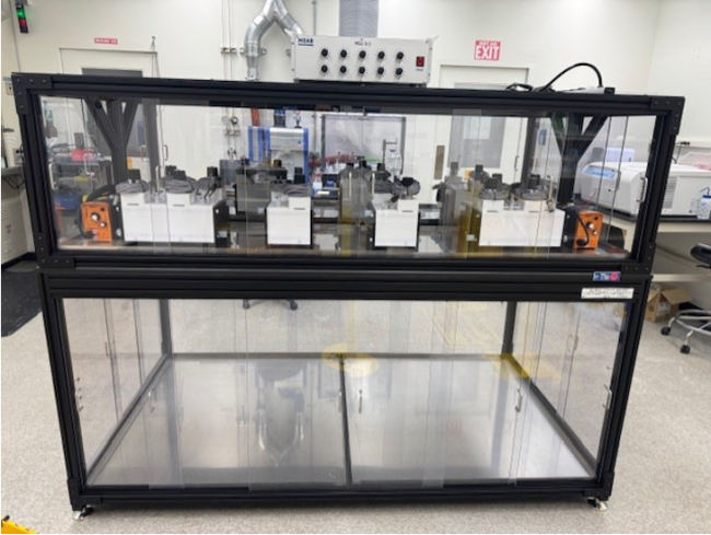 Image of the mixer settler system installed in a ventilated enclosure within the radiological facility. The controllers are placed on top of the enclosure, and liquid feed/collection containers will be positioned beneath the work surface. The mixer settler units are accessible from all four sides via sliding doors.